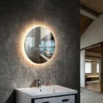 A round backlit mirror casts a warm glow in a modern bathroom above a sleek white vanity and sink