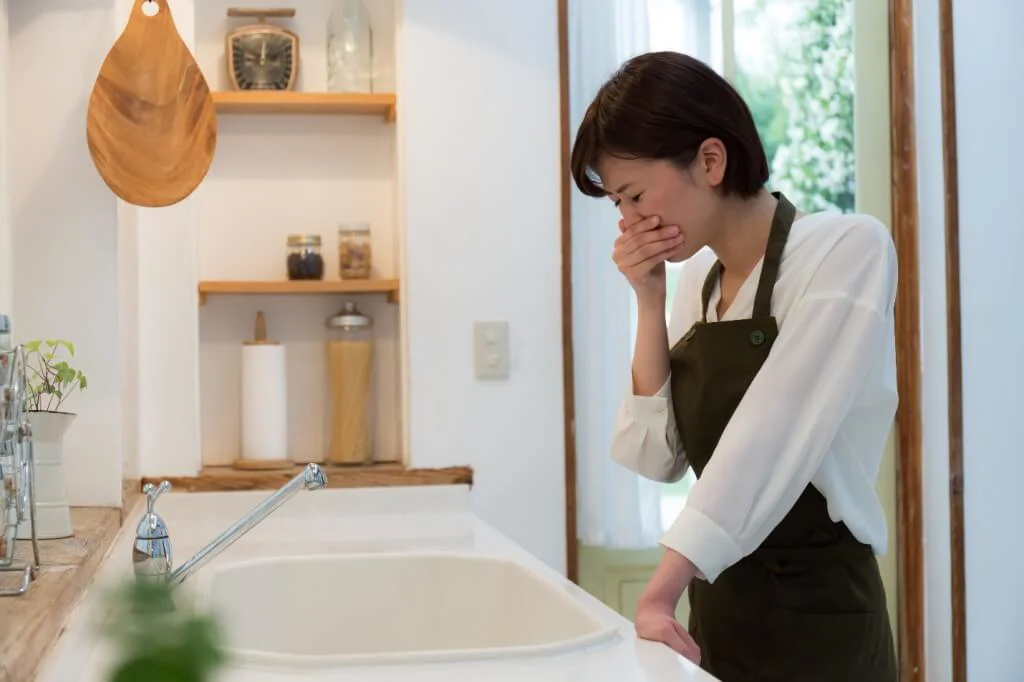 A woman in a kitchen looks distressed, holding her hand over her mouth near a white sink, appearing nauseated by a foul odor from a clogged drain