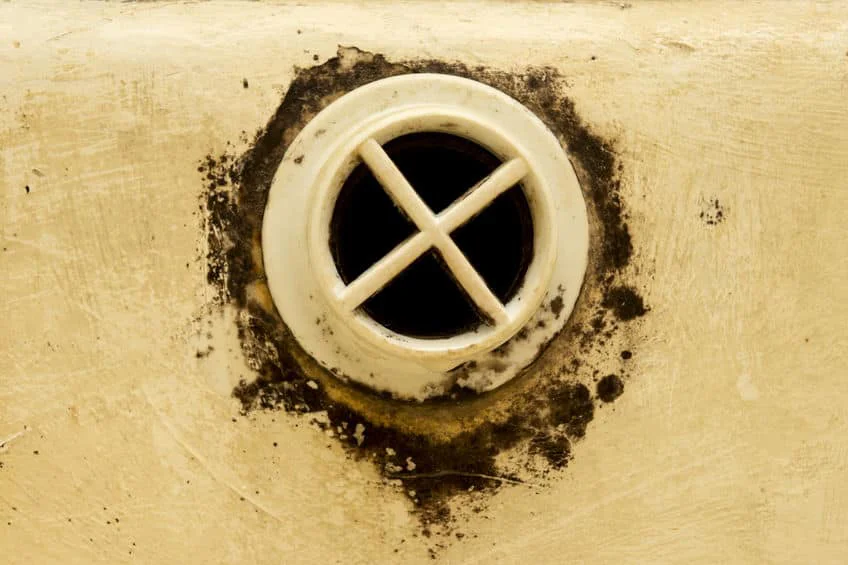 A grimy, white overflow drain with a cross-shaped grate is seen on a light-colored surface