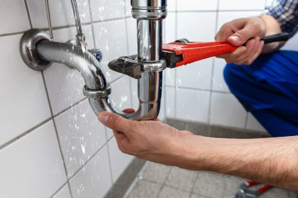 A plumber uses a red wrench to tighten a leaking chrome P-trap pipe under a sink