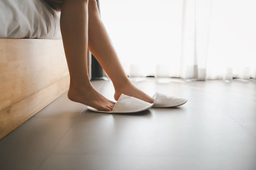Bare feet sit at the edge of a bed, reaching for white slippers on a bright, tiled floor