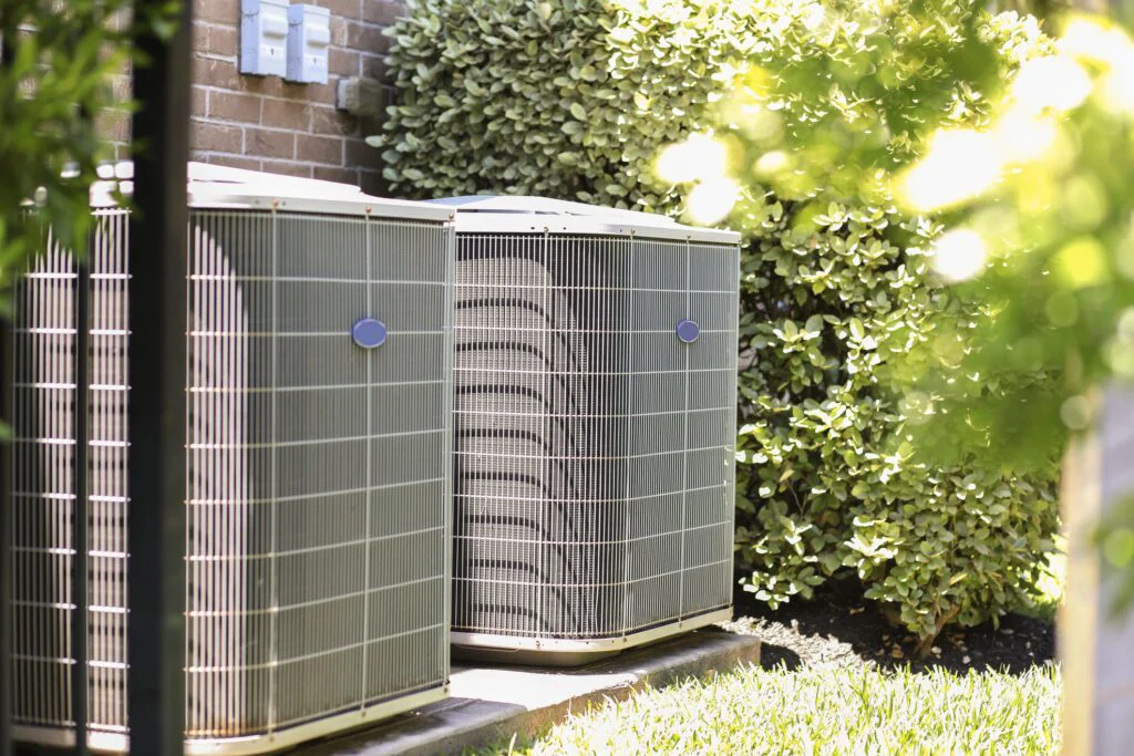 Two outdoor HVAC condenser units stand against a brick wall, surrounded by green landscaping