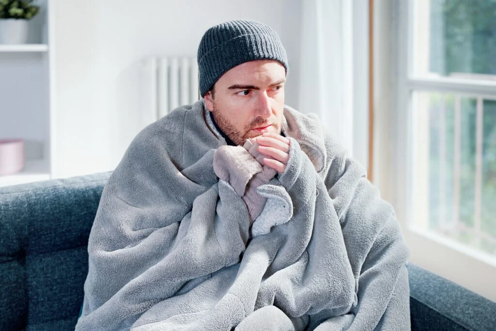 A man bundled in a gray blanket and beanie looks cold and concerned while sitting indoor