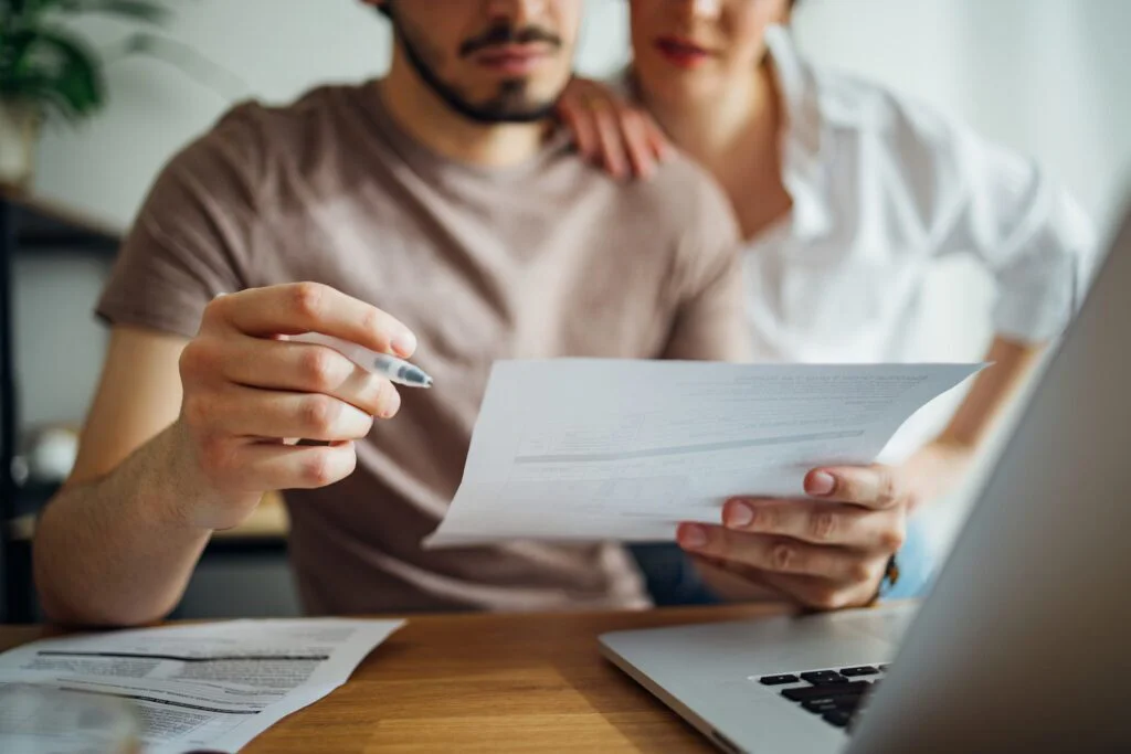 A couple sits at a wooden table, reviewing home financial documents with a laptop open