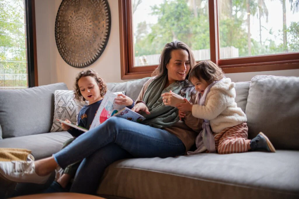 A mother and her two children enjoy reading a book together on a sofa, creating a warm and secure family scene in their home