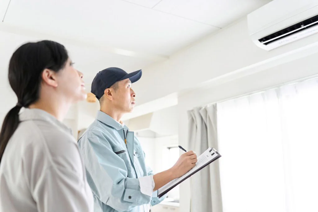 A technician in uniform holds a clipboard, inspecting a wall-mounted air conditioner