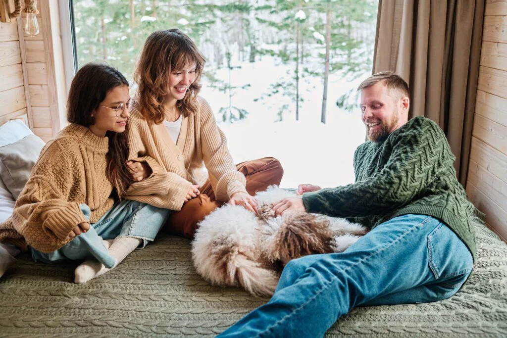 A happy family, with two women, a man, and a dog, relaxes by a large window overlooking a snowy forest