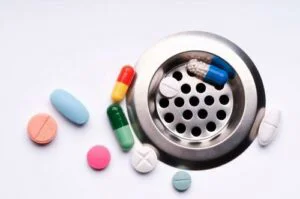 Colorful pills are scattered near and in a sink drain, representing improper medication disposal