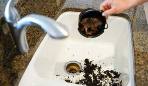 A person is seen dumping wet coffee grounds from a filter basket into a white kitchen sink